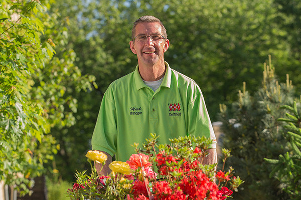 Mark, a garden center manager, in his green uniform, rolling a cart of roses and flowers through the nursery yard.